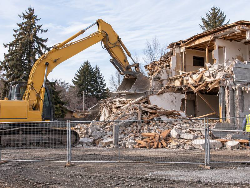 Excavator beside a partly demolished house inside a temporary site fence